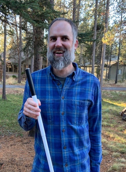 A white male wearing a blue plaid button up shirt, holding a cane for the blind, is smiling at the camera, with trees and cabins in the background.